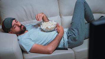 Tired man relaxing on sofa in front of television eating popcorn while watching movie show. Caucasian male with beard looking entertainment series late at night in kitchen photo
