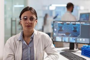 Scientist with lab coat sitting in laboratory looking at camera smiling, while colleague working on computer in background. Chemist examining virus evolution using high tech for scientific research, vaccine. photo