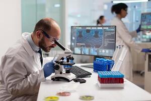 Microbiologist working to find a vaccine for virus in lab looking at sample through microscope weaing protection glasses. Specialists working on medicine, biotechnology research in advanced pharma lab. photo