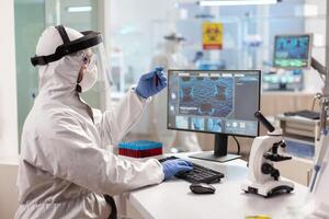 Scientist dressed in ppe suit working on computer holding vaccutainer. Medical team examining vaccine evolution in medical lab using high tech and chemistry tools for scientific research photo