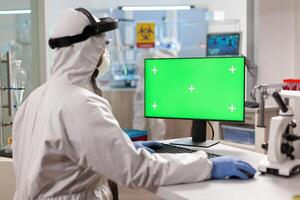 Man researcher in coverall looking at chroma key computer in modern equipped lab. Team of microbiologists doing vaccine research writing on device with chroma key, isolated display. photo