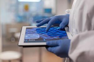 Close up of chemist in ppe suit using tablet pc to check virus vaccine. Scientist examining virus evolution using high tech technology researching vaccine development against covid19. photo