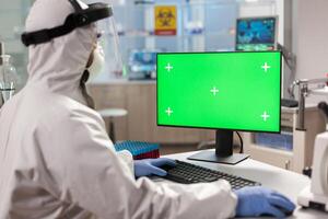 Scientist man wearing protection suit typing on computer with green mockup. Team of microbiologists doing vaccine research writing on device with chroma key, isolated display. photo