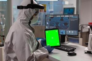 Doctor with coverall working on tablet with green screen in modern equipped lab.Team of microbiologists doing vaccine research writing on device with chroma key, isolated display. photo