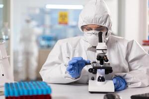 Healthcare scientist dressed in ppe suit working using microscope in modern lab. Chemist in coverall working with various bacteria, tissue blood samples for antibiotics research. photo