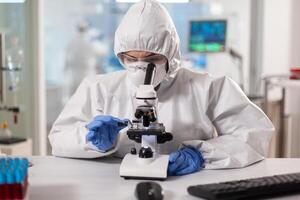 Scientist in ppe suit making adjustments and looking through laboratory microscope. Chemist in coverall working with various bacteria, tissue blood samples for antibiotics research. photo