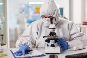 Scientist working to discover virus vaccine dressed in ppe suit looking through microscope. Chemist in coverall working with various bacteria, tissue blood samples for antibiotics research. photo
