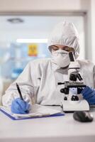 Doctor writing notes dressed in ppe suit and visor mask using microscope. Chemist in coverall working with various bacteria, tissue blood samples for antibiotics research. photo