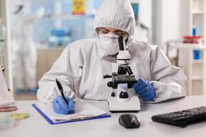 Medicine scientist dressed in ppe suit taking notes during vaccine research using microscope. Chemist in coverall working with various bacteria, tissue blood samples for antibiotics research. photo