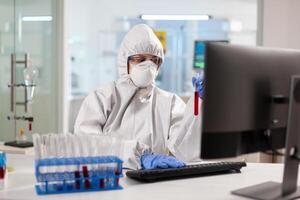 Medical researcher dressed in ppe suit looking at blood sample using computer. Scientist examining vaccine evolution using high tech technology for treatment development against covid19 virus. photo