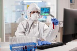 Researcher in coverall holding test tubes with blood sample typing on computer. Scientist examining vaccine evolution using high tech technology for treatment development against covid19 virus. photo