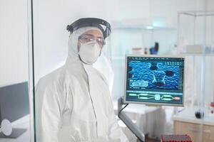 Tired doctor in protection suit against covid-19 looking exhausted at camera behind the glass wall working in danger area. Scientist examining virus evolution using high tech for scientific research photo