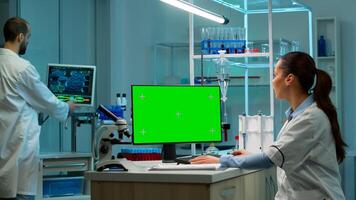 Biochemist sitting at workplace in laboratory using green mock-up screen personal computer with chroma key monitor. coworker working in background of pharmaceutical research centre. photo