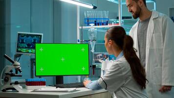 Female scientist working on computer with mock-up green screen in modern laboratory. Scientist doctor using various shelves with beakers, chemicals and different technical equipment is visible. photo