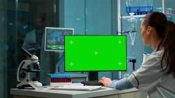 Over the shoulder shot of chemist working with green screen mock-up template on desktop computer, isolated display, chroma key. In background scietist doctor enter in medical lab with blood sample. photo