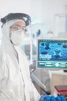 Microbiologist in ppe suit standing in laboratory looking at camera behind the glass wall in equipped lab. Doctor examining virus evolution using high tech and chemistry tools for vaccine development. photo
