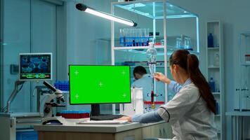 Microbiologist working on computer with display, green chroma key screen talking with coworker about new vacutainer with blood samples. Team of biotechnology scientists developing drugs. photo