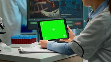 Back view of chemist woman holding tablet with green mockup in modern equipped lab. Team of microbiologists doing vaccine research writing on device with chroma key, isolated display. photo