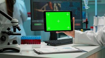 Close up of scientist man holding tablet with green mockup in modern equipped lab. Team of microbiologists doing vaccine research writing on device with chroma key, isolated display. photo