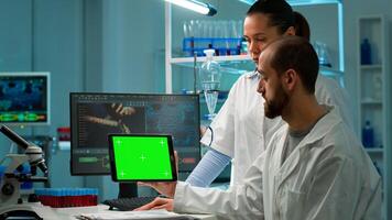 Man research scientist has conversation with famale bioengineer next to a desktop computer holding tablet with green screen mock up. They look at chroma key pc template in a modern science laboratory. photo