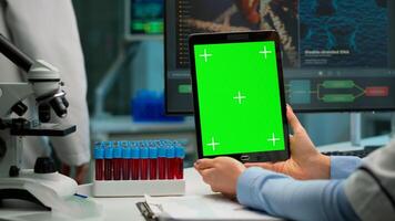Close up of scientist woman holding tablet with green mockup in modern equipped lab. Team of microbiologists doing vaccine research writing on device with chroma key, isolated display. photo