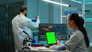 Microbiologist working on notepad with green chroma key display in modern equipped lab. Team of biotechnology scientists developing drugs using tablet with mock up screen. photo