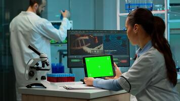 Biochemist sitting at workplace in laboratory using green mock-up screen tablet with chroma key display while coworker working in background of pharmaceutical research centre, bringing blood sample photo