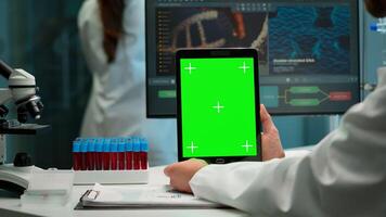 Close up of biochemist sitting at workplace in laboratory using green mock-up screen tablet with chroma key display. coworker working in background bringing blood sample. photo