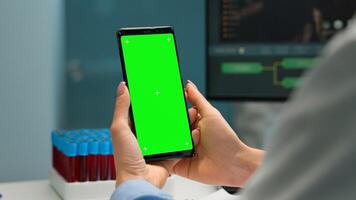 Close up of doctor's hand holding phone with green screen sitting at desk in biological laboratory while nurse bringing blood samples. Scientist using smartphone with mockup, chroma key display photo