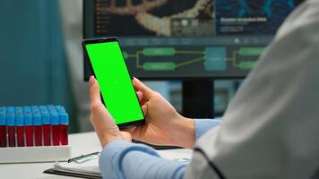 Medic in scientific laboratory holding phone with greenscreen wearing white coat while nurse bringing blood samples. Healthcare specialist in modern lab using smartphone with mockup chroma key display photo