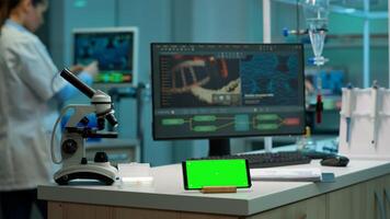 Scientist standing at monitor in background analysing virus evolution while phone with mock-up green screen working in front. Woman lab researcher examining vaccine developent bringing blood samples photo
