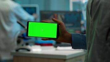 Microbiologist holding smartphone with green chroma key display in front of camera in modern equipped lab. Team of biotechnology scientists developing drugs using tablet with mock up screen. photo