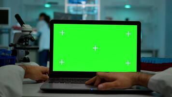 Pov shot of man scientist sitting at desk working on laptop with mock-up green screen, isolated display. In background lab researcher analysing vaccine developent examining samples photo