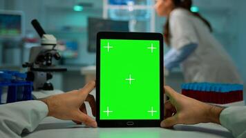 Pov shot of man scientist sitting at desk working on notebook with mock-up green screen, isolated display. In background lab researcher analysing vaccine developent examining samples photo