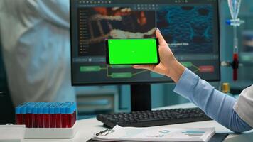 Close up of scientist woman holding smartphone with green mockup in modern equipped lab. Team of microbiologists doing vaccine research writing on device with chroma key, isolated display. photo