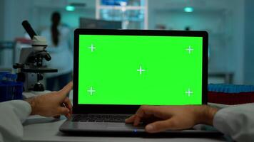 Pov shot of microbiologist typing on laptop with green chroma key display sitting at desk working reading virus symptoms. In background lab researcher analysing vaccine developent examining samples photo