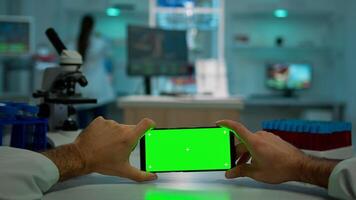 POV shot of chemist using horizontal smartphone with chroma key on isolated display in biological laboratory. Doctor wearing white coat in clinic working with mobile with green screen in medical lab photo