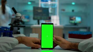 Pov shot of microbiologist holding phone with green chroma key display sitting at desk searching, reading virus symptoms. In background lab researcher analysing vaccine developent examining samples photo