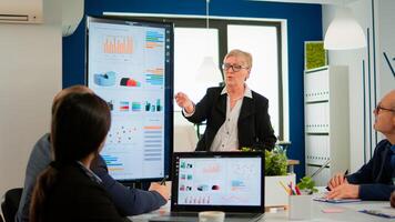 Elderly project manager pointing at desktop presenting statistical data, briefing diverse group of employees. Multiethnical team working in professional startup financial company during conference photo