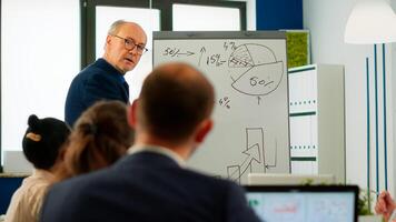 Elderly businessman giving presentation to multiethnic team of colleagues using flip chart, drawing, explaning financial graphs in meeting room. Manager interacting with audience at corporate workshop photo