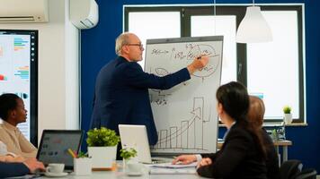 Senior businessman in eyeglasses and formal suit drawing financial plan on flipchart and explaining to interracial team of colleagues while giving presentation during conference meeting, brainstorming photo