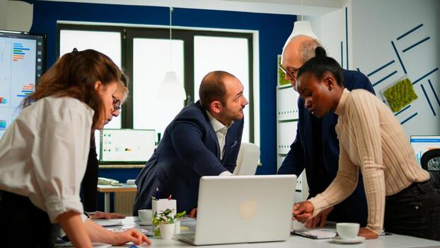Diverse businesspeople working in teamwork discussing project strategy using laptop standing at conference table. Manager executive analysing financial data during business meeting in broadroom photo