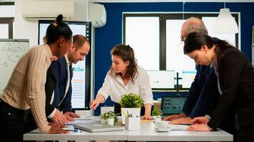 Diverse team of workers talking, looking at documents and analyzing graphs data standing at conference desk in start up company. Professional colleagues team discussing marketing project brainstorming photo