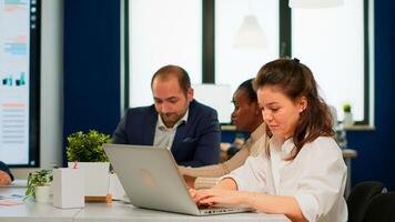 Satisfied business woman answering emails typing on laptop smiling sitting at desk in busy start up office while diverse team analyzes statistics data. Multiethnic team working at new project photo