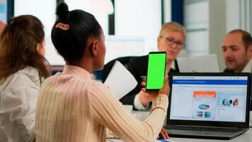 Back view of black businesswoman sitting at conference desk holding smartphone with green screen while colleagues working on background. Multiethnic coworkers planning project on chroma key display photo