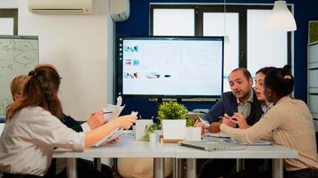 Executive explaining company's vision and potential to employees sitting at brainstorming table in broadroom with TV screen on the wall showing corporate growth photo