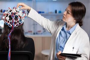 Back view of woman patient wearing performant eeg headset sitting on chair in neurological research laboratory while medical researcher adjusting it, examining nervous system typing on tablet. photo