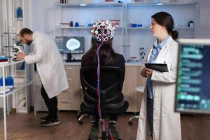 Back view of woman wearing headset for brain scan in neurology laboratory, neuro science. Nervous system analysed by team of scientists. Medical scientist looking through microscope in background developing treatment. photo