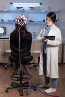 Woman patient wearing performant eeg headset scanning brain electrical activity in neurological research laboratory while medical researcher adjusting it, examining nervous system typing on tablet. photo