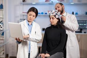 Specialist neurologist doctor taking notes on laptop asking patient's symptoms adjusting high tech eeg headset. Doctor researcher controlling EEG headset analyzing brain functions and health status. photo